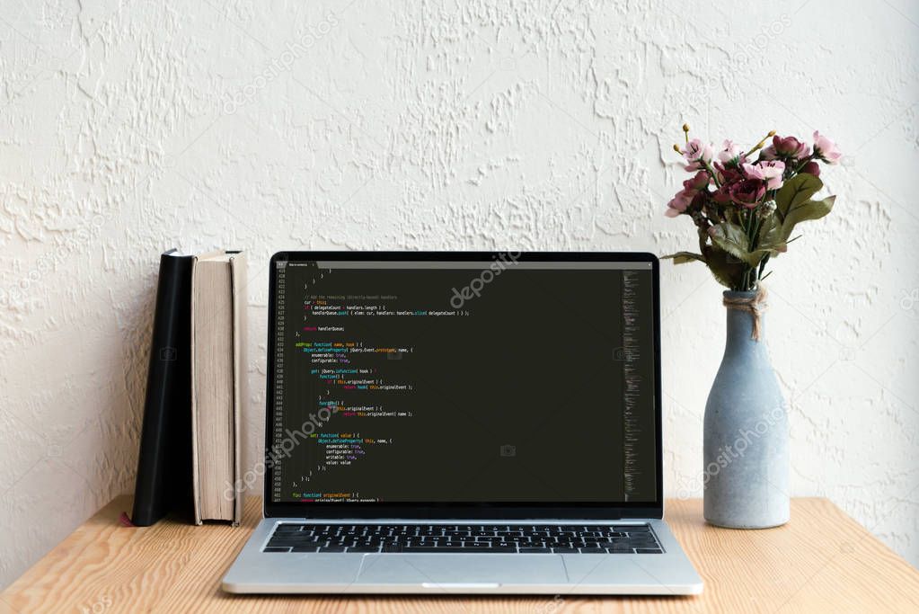 laptop with html code on screen, books and flowers in vase on wooden table